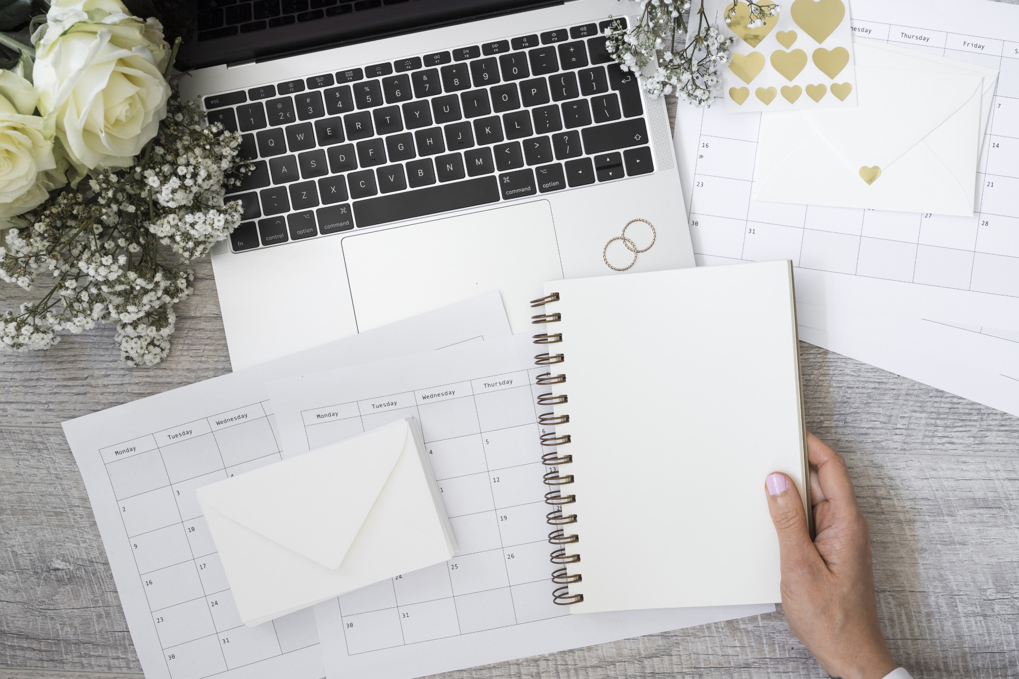 close up of a person holding blank spiral notebook with laptop wedding rings flower envelope and calendars on wooden desk